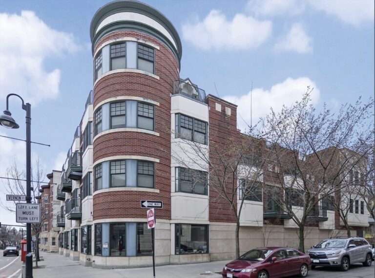 Corner brick residential building with rounded turret, large windows, and balconies on a city street, blue sky above.